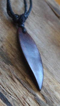 Load image into Gallery viewer, A dark wooden Surferkette Holz Halskette with leaf-shaped pendant and adjustable cotton cord rests on a rustic wooden surface.
