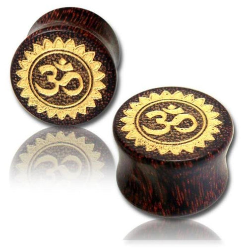 Two round Holz Plug Tamarind Wood Tribal Tunnels feature hand-carved and gold-painted Om designs with a laser-engraved sunburst, displayed on a white background with subtle reflection.