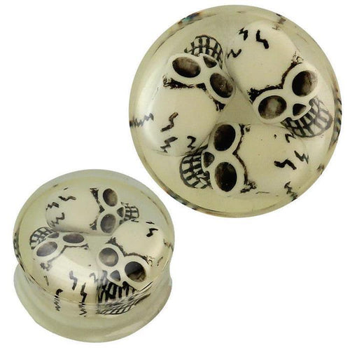 A transparent acrylic plug featuring three black and white 3D Totenkopf skulls, glowing in the dark. The floating skull design creates a striking effect for piercing and ear jewelry enthusiasts.