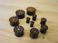 Load image into Gallery viewer, Nine Plug Arangholz Wikinger Kompass Inlay double flared plugs of various sizes, featuring rune-inspired circular carvings, are displayed on a light wood surface, each piece showcasing a decorative, mystical Norse design.
