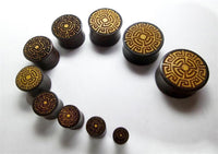 Load image into Gallery viewer, Ten "Plug Tamarindenholz griechisch Sonne Ornament Spiralen Inlay double flared flach" plugs, each round and intricately inlaid with golden sun spiral patterns, are displayed in a spiral from smallest to largest on a white background.
