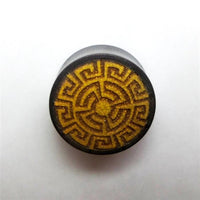 Load image into Gallery viewer, A Plug Tamarindenholz griechisch Sonne Ornament Spiralen Inlay double flared flach with a yellow and black geometric sun pattern and central dot, made of tamarind wood, is shown on a white background.

