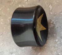 Load image into Gallery viewer, A black Horn Plug Stern Inlay metallic gold Flesh Tunnel Expander, hand-crafted, with a shiny surface and gold star detail on one end, rests on a textured light gray surface.
