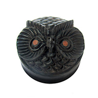 Load image into Gallery viewer, The "Holz Plug Eule schwarz handgeschnitzt braune Augen" is a round, dark wood plug with an owl’s face, detailed feathers, brown eyes, and a small beak—an eye-catching organic Schmuck shown on a white background.
