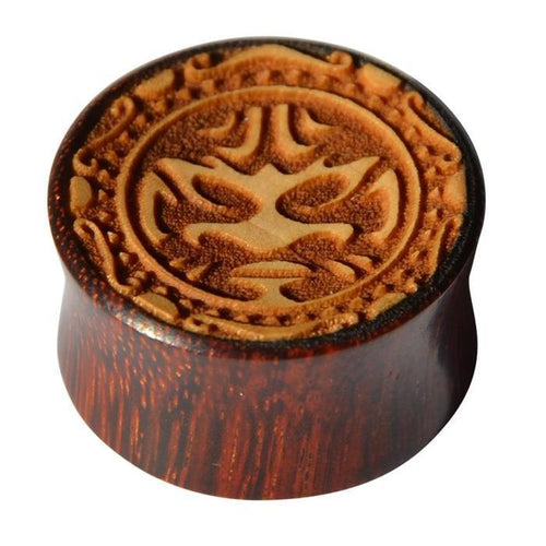 The Plug Holz hell dunkel Tribal Maske Gravur double flared is a round tamarind wood plug with a polished reddish-brown finish, featuring a carved tribal mask design surrounded by intricate decorative patterns.