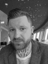 Load image into Gallery viewer, A tattooed, bearded man with a nose ring and ear gauges—one showcasing the Plug Celtic Sawoholz Holz braun Geflochten Lasergravur—sits indoors by large windows under a ceiling dotted with star-like lights.
