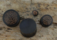 Load image into Gallery viewer, Four round, dark Holz Plug Kokosholz Inlay handgeschnitzt Sonoholz plugs with brown, speckled tops are arranged on a rough, light brown wooden surface. Their unique spotted pattern and size variation highlight their handmade quality.
