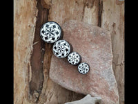 Load image into Gallery viewer, Four round objects, including a "Plug Horn weiß Gravur schwarz Bänder Blende Sternmuster" featuring a black and white star pattern, are arranged in a row from largest to smallest on textured stone with a wooden background.

