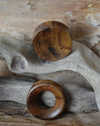 Load image into Gallery viewer, A Flesh Plug Holz Parasitholz braun double flared—one solid, one with a circular hole—is displayed on driftwood against a rustic wooden background.
