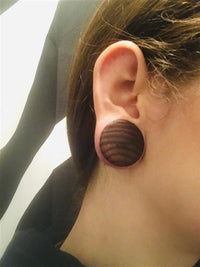 Load image into Gallery viewer, Close-up of a person’s ear wearing the hand-carved Zebraholz Plug "Holzplug Zebraholz handgeschnitzt braun gestreift Tribal Holz Tunnel Plug Expander" with brown stripes, stretching the earlobe. Background: black and white fabric.
