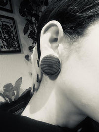 Load image into Gallery viewer, A close-up black-and-white photo shows a person wearing a large, round Holzplug Zebraholz handgeschnitzt braun gestreift Tribal Holz Tunnel Plug Expander. Their hand touches their neck, with blurred leafy plants and framed art in the background.
