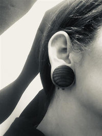 Load image into Gallery viewer, Close-up black and white photo of an ear with a large, dark Holzplug Zebraholz handgeschnitzt braun gestreift Tribal Holz Tunnel Plug Expander. Hair is tucked behind the ear, striped background mirrors the plug’s unique zebrawood pattern.
