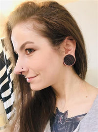 Load image into Gallery viewer, A woman with long brown hair and a nose piercing smiles slightly, wearing tribal zebrawood hand-carved plugs (Holzplug Zebraholz braun gestreift), standing indoors near a striped shower curtain. A cat tattoo is visible on her chest.
