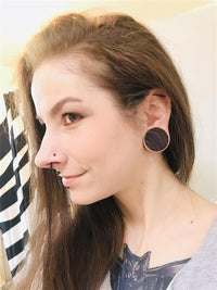 Load image into Gallery viewer, A person with long brown hair and stretched earlobes wears large brown striped Holzplug Zebraholz handgeschnitzt Tribal Holz Tunnel Plugs, a nose piercing, and a cat chest tattoo. Shown in profile, they smile slightly.
