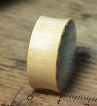Load image into Gallery viewer, A close-up of the Plug Krokodilholz crushed Türkis Sonne double flared, showing its smooth light-colored side and patterned top with blue and gray Türkis inlay, resting on wood beside a ruler.
