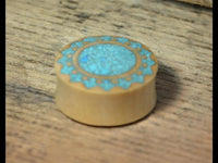 Load image into Gallery viewer, A round, light-colored Plug Krokodilholz crushed Türkis Sonne double flared rests on textured wood. The top displays a blue mosaic reminiscent of Türkis inlay within a circular border.
