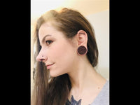 Load image into Gallery viewer, A woman with long brown hair and a chest tattoo is shown in profile, wearing a large Herz Blumen Muster Lasergravur Sawoholz Tribal Plug in her stretched ear lobe as she gazes slightly forward.
