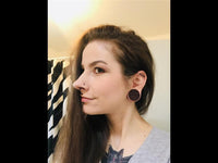 Load image into Gallery viewer, A woman with long brown hair, a nose stud, wearing large round Herz Blumen Muster Lasergravur Sawoholz Tribal Plugs and a cat tattoo on her chest, smiles slightly as she poses for an indoor photo.
