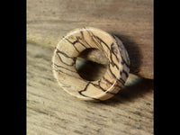 Load image into Gallery viewer, The Bananenholz Flesh Tunnel hell ribbed straight konkav features a polished, round design with a central hole and natural dark grain patterns, evoking the look of a Holztunnel, shown resting on textured wood.
