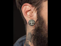 Load image into Gallery viewer, A close-up of a bearded person with a neck tattoo wearing the Holz Plug Baum des Lebens Yggdrasil silber double flared earring, featuring a detailed tree design, set against a dark background.
