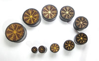 Load image into Gallery viewer, Nine round black Plug Arangholz Wikinger Kompass Inlay double flared plugs in various sizes are arranged in a curve on a white background, each with a gold Viking rune symbol for a unique and bold set.
