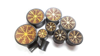 Load image into Gallery viewer, Seven round, black Plug Arangholz Wikinger Kompass Inlay double flared plugs with brown tops feature gold Vegvisir rune designs on a white background. One plug is tipped forward, revealing its side profile.
