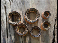 Load image into Gallery viewer, Six handmade Holz Plug Olivenholz Sonnenblume double flared plugs with intricate black geometric patterns along their edges are arranged on a weathered wooden surface. The plugs vary in size.
