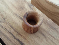 Load image into Gallery viewer, A small, smooth Olivenholz Flesh Tunnel Ohr Piercing Holz Double Flared rests on wood with a matching grain and color, creating a natural, seamless look.
