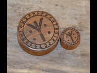 Load image into Gallery viewer, Two round Holz Plug Teakholz Rune Leben Tod Punkte double flared tokens with engraved runic symbols rest on wood. The larger plug shows a bold Rune Algiz at its center, surrounded by smaller runes.
