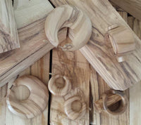 Load image into Gallery viewer, The Plug Olivenholz Pin-Creole braun gemasert Holz-Pin Creole, featuring natural light brown wood grain, rests on wooden planks alongside other wooden rings and jewelry pieces in round and angular shapes.
