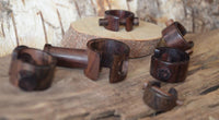 Load image into Gallery viewer, The Plug Narraholz Pin-Creole braun gemasert Holz-Pin Creole, featuring unique geometric designs, is displayed on a rustic wooden surface and a small log, set against a textured wood backdrop.
