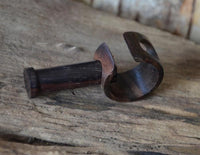Load image into Gallery viewer, The Plug Narraholz Pin-Creole braun gemasert Holz-Pin Creole, featuring a dark, polished finish and artisanal design, rests on a rustic wooden surface with its cylindrical handle and curved, semi-enclosed blade made from Narraholz.
