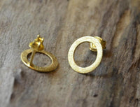 Load image into Gallery viewer, The "Ohrstecker Silber 925 vergoldet Creolen Ringe gebürstet" are small, matte-finish stud earrings. One earring shows its open circle design facing up, while the other lies on its side on a wooden surface.
