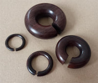 Load image into Gallery viewer, Four dark brown Piercing Narraholz Holz Expander rings in different sizes are displayed on a beige surface; three rings feature a small gap, and the largest Holz Expander has a wider opening.

