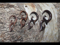 Load image into Gallery viewer, Four Sonoholz lang Tribal Piercing Holz Bögen Ornament earrings, showcasing hand-carved craftsmanship, are displayed on textured tree bark in a row, arranged from left to right by increasing size and darkness.
