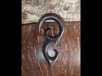 Load image into Gallery viewer, The Sonoholz lang Tribal Piercing Holz Bögen Ornament features curved, flowing shapes carved from wood, displayed on a smooth brown surface with a textured, light-colored backdrop above.
