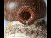 Load image into Gallery viewer, The Ohr Donut Piercing Dahlie Holz Ohrgewicht Ring is a circular wooden hair accessory with a geometric pattern, featuring a small bottom opening, displayed on textured bark with a coconut shell in the background.
