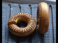 Load image into Gallery viewer, The Ohr Donut Piercing griechisch Jackfrucht Holz features two circular wooden rings with a Greek key pattern, each having a small side opening. One ring lies flat while the other stands upright.
