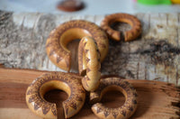 Load image into Gallery viewer, Five Ohr Donut Piercing Pfau Mandala rings made from Jackfrucht Holz with intricate burned designs rest on a bark piece. The light brown wooden jewelry features dark mandala patterns, small holes, and are arranged overlapping and leaning.
