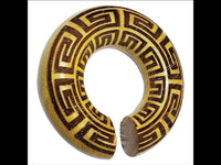 Load image into Gallery viewer, The Ohr Donut Piercing griechisch Jackfrucht Holz is a round, hollow wooden bangle with a small opening, adorned with a yellow and brown Greek key pattern, displayed against a white background and resembling an elegant donut accessory.
