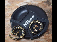 Load image into Gallery viewer, A pair of Brass Piercing Dehnschnecke Resin schwarz gold Spiralen earrings rests on a black Nikon camera lens cap, set on a wood-textured surface.
