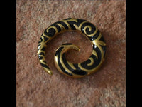 Load image into Gallery viewer, A Brass Piercing Dehnschnecke Resin schwarz gold Spiralen earring featuring ornate black and gold patterns rests on a textured, reddish-brown stone surface.

