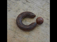 Load image into Gallery viewer, The Captive Bead Klemmkugel Ring Sawoholz Piercing Mandala is a crescent-shaped, brown wooden ring with intricate carvings and a smooth round bead on one end, shown here resting on a light wooden surface.
