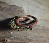 Load image into Gallery viewer, The Piercing Dehnschnecke Narraholz Brass Ornament Spirale is a wooden spiral earring with brass detailing, displayed on a textured wooden surface in soft light—ideal for lobe piercings.
