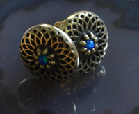 Load image into Gallery viewer, A pair of Messing Stein Ohrstecker antik gold Dahlie 10mm earrings features vintage-style round studs with intricate lattice and blue-green opal-like centers, reminiscent of a dahlia flower, shown on a dark reflective surface.
