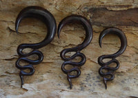 Load image into Gallery viewer, Three dark brown, intricately twisted Holz Lobe Piercing Dehner hooks are displayed on textured light-brown stone—perfect for lobe piercing enthusiasts.
