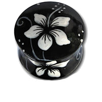 Load image into Gallery viewer, The Holz Plug Hawaii Blume weiß schwarz Teakholz handbemalt is a small, round black plug with a lid, featuring a hand-painted white flower design—ideal as unique jewelry or to pair with your Hibiskus Piercing.

