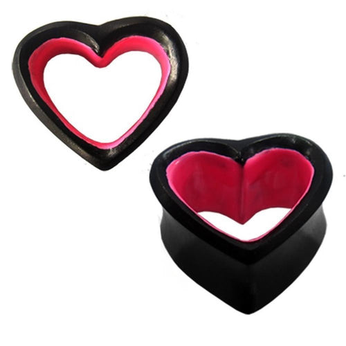 Two hand-carved Sonoholz Holztunnel Herz plugs, in black with pink inner edges, display their hollow heart design—one upright, one on its side—highlighting the unique tribal style of these ear plugs.