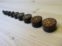 Load image into Gallery viewer, A row of Plug Arangholz Wikinger Kompass Inlay double flared plugs, engraved with yellow Norse runes including a Vegvisir, are arranged from largest to smallest on a light wooden surface.
