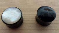 Cargar imagen en el visor de la galería, Two Buffalo Horn Plug Perlmutt Inlay weiß handgeschnitzt Lobe Piercing Tunnels rest on wood: the left with a shiny white Perlmutt inlay, the right solid black—ideal organic expanders for stretched piercings.
